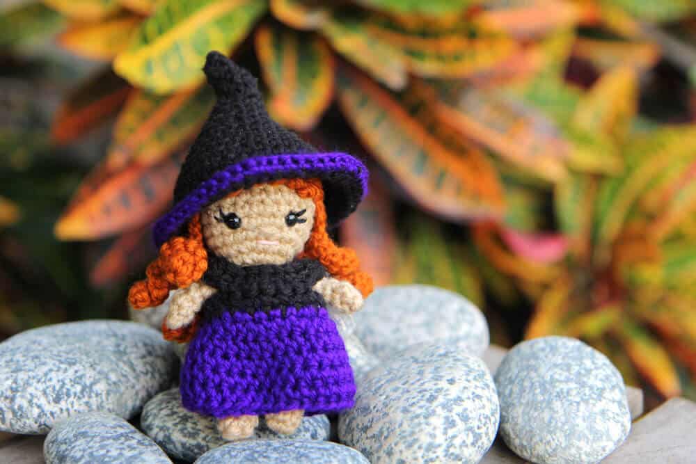 Tiny crochet witch with a purple and black dress and a black witch hat accented with a purple brim