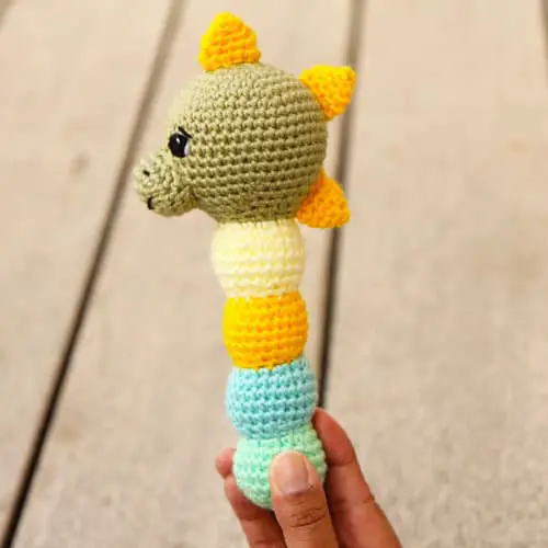 dinosaur amigurumi baby rattle with a green head, yellow spikes and a yellow blue and green stem