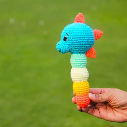 crochet dinosaur baby rattle with blue head, coral spikes, and a multi-coloured stem