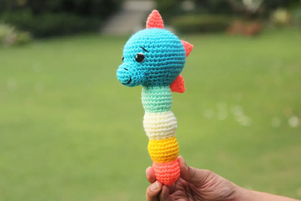 A crocheted dinosaur baby rattle with a blue head, coral colours spikes, and a stem made up of four multicoloured balls for an easy grip