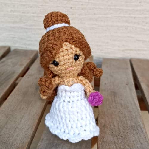 Crochet bride doll with a white dress with a sweetheart neckline. Half her hair is up in a bun and the rest is in curls around her shoulder. She is carrying a purple bouquet